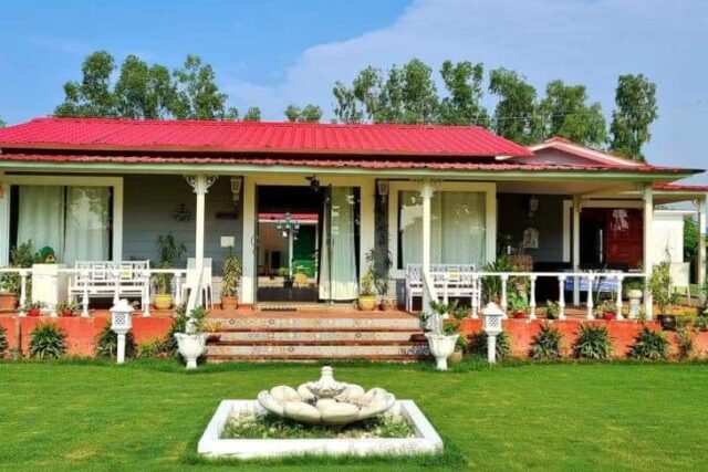 A green beauty farmhouse located in Noida Sector 135 with a vibrant red roof, surrounded by lush greenery and a manicured lawn. The property features a spacious front porch with potted plants, a decorative water fountain, and modern amenities. Solar panels and surrounding farms add to the sustainable charm.