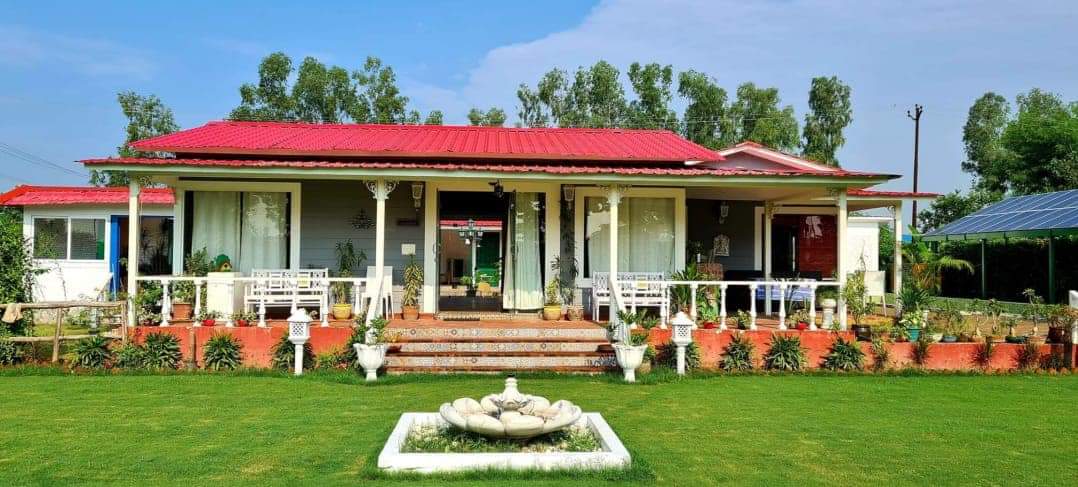 A green beauty farmhouse located in Noida Sector 135 with a vibrant red roof, surrounded by lush greenery and a manicured lawn. The property features a spacious front porch with potted plants, a decorative water fountain, and modern amenities. Solar panels and surrounding farms add to the sustainable charm.